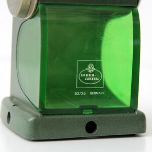 Pu&ograve; includere: Un temperamatite Faber-Castell verde con un contenitore per trucioli in plastica verde traslucida. Il contenitore presenta il logo bianco Faber-Castell, il testo 52/25 e la parola GERMANY stampata sul davanti. La base &egrave; verde scuro.