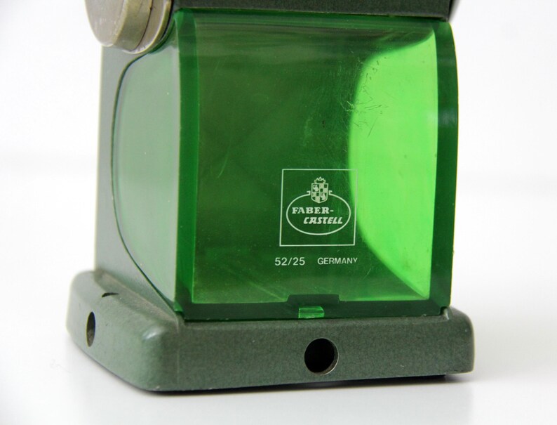 Pu&ograve; includere: Un temperamatite Faber-Castell verde con un contenitore per trucioli in plastica verde traslucida. Il contenitore presenta il logo bianco Faber-Castell, il testo 52/25 e la parola GERMANY stampata sul davanti. La base &egrave; verde scuro.