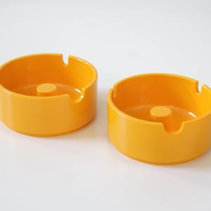 May include: Two round, bright yellow ashtrays. Each ashtray has a cylindrical shape with a central post and a notch for resting cigarettes. The ashtrays are made of a smooth, glossy material.