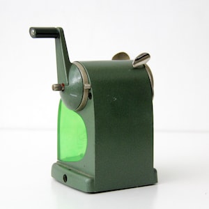 Pu&ograve; includere: Un temperamatite manuale vintage con corpo in metallo testurizzato verde oliva, manovella in plastica nera e un contenitore per i trucioli in plastica verde brillante traslucida. Il dispositivo include una leva superiore in metallo argentato e una piastra frontale.