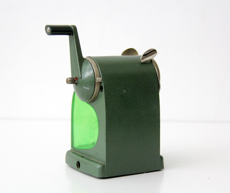 Pu&ograve; includere: Un temperamatite manuale vintage con corpo in metallo testurizzato verde oliva, manovella in plastica nera e un contenitore per i trucioli in plastica verde brillante traslucida. Il dispositivo include una leva superiore in metallo argentato e una piastra frontale.