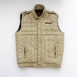 Könnte beinhalten: Beige gesteppte Weste mit hohem Kragen und zwei Vordertaschen. Die Weste hat ein braunes Innenfutter und den Text "Lord Anthony" auf einem kleinen Etikett. Schulterklappen und gerippter Saum.