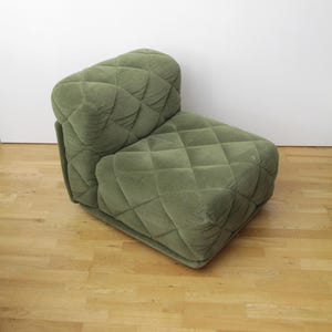 May include: A low-profile, armless lounge chair upholstered in a sage green fabric with a prominent diamond-quilted pattern. The chair features a thick, cushioned seat and a backrest that curves slightly, creating a soft, rounded silhouette.