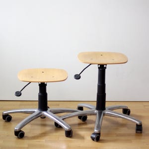 May include: Two adjustable stools with light wood seats and black central columns. Each stool features a five-point silver metal base with black casters. A black lever is attached under each seat for height adjustment, showing two different heights.