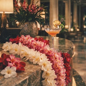 May include: A cocktail in a stemmed glass garnished with a pineapple slice and lime peel sits on a marble counter. A lei of red, pink, and white flowers adorns the counter. A lamp and floral arrangement are in the background.