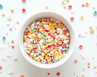 May include: A white bowl filled with a colorful mix of breakfast cereal, featuring small red, blue, yellow, and green ring-shaped pieces combined with white puffed grains, scattered across a plain white surface.