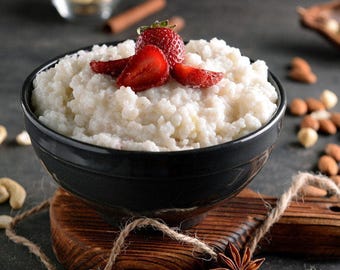 May include: A dark gray bowl filled with white rice pudding topped with fresh red strawberry slices. The bowl rests on a wooden board tied with twine, accompanied by scattered nuts, a cinnamon stick, and a star anise spice.