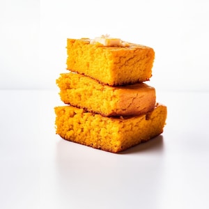 May include: A stack of three golden-yellow cornbread squares, each with a textured surface. The top piece is topped with a small pat of butter. The cornbread is set against a bright white background.