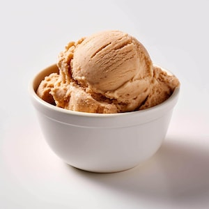 May include: A white ceramic bowl filled with scoops of light brown ice cream. The ice cream has a textured surface and appears to be freshly scooped. The bowl is round and sits on a white surface.