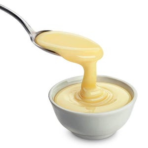 May include: A silver spoon pouring a stream of thick, pale yellow liquid into a small, white bowl. The bowl is filled with the same creamy substance. The background is plain white.