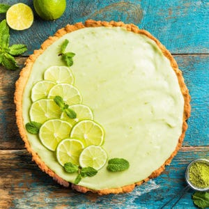 May include: A key lime pie with a light green filling, garnished with lime slices and mint leaves. The pie sits on a golden-brown crust, with a rustic wooden background. A small bowl of green powder is in the corner.
