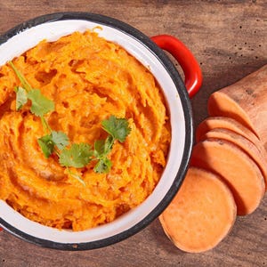 May include: Overhead shot of a bowl of orange mashed sweet potatoes garnished with cilantro. Sliced sweet potatoes and a whole sweet potato are on a wooden surface. The bowl has red handles.