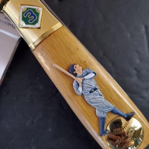 May include: A baseball bat-shaped letter opener with a wooden handle and gold accents. A baseball player figurine in a blue and white uniform is featured on the handle. A baseball and the number 3 are also visible.