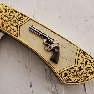 May include: A decorative pocket knife with a gold-colored filigree design on the edges. The knife features a silver-colored revolver and a light-colored wood grain center.