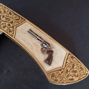 May include: A folding knife handle featuring a cream-colored center inlay with a small silver-toned revolver emblem. The handle is framed by gold-toned metal ends decorated with intricate, embossed floral scrollwork engravings.