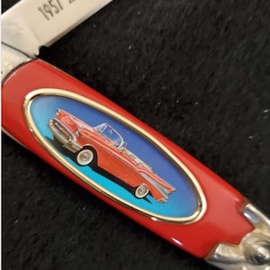 May include: A red pocket knife with a silver blade and accents. The handle features an oval design with a red convertible car on a blue background, framed in gold. The blade reads "1957 Bel Air".