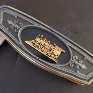May include: A decorative, gray and gold-toned metal belt buckle featuring a gold-colored steam train design on a black oval. The buckle has a decorative border and fleur-de-lis accents.