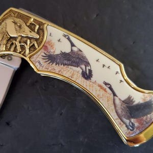 May include: A folding pocket knife with a decorative handle featuring a gold-colored goose head and a painted scene of geese in flight. The handle has a cream-colored background with gold trim.