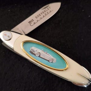 May include: A vintage pocket knife with a cream-colored handle and a blade engraved with "1953 Chevrolet Corvette". The handle features a car graphic on a teal background, framed in gold. A small compass is on the handle.