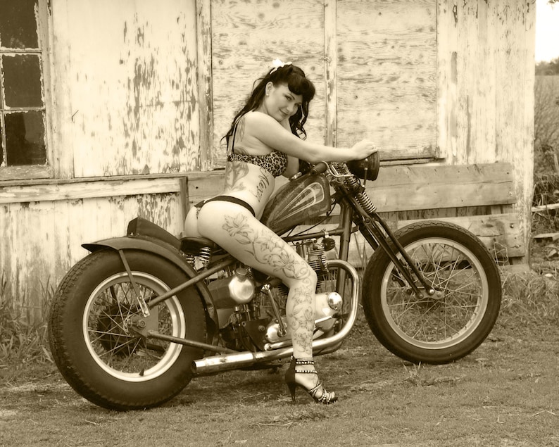 May include: A woman in a bikini sits on a vintage motorcycle with a black leather seat. The motorcycle has a chrome exhaust pipe and a black fender. The woman has tattoos on her legs and is wearing a black bikini bottom and a leopard print bikini top. The motorcycle is parked in front of a weathered wooden building.