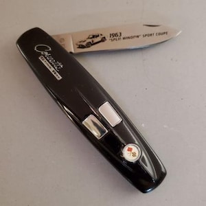 May include: A black pocket knife with the word "Corvette" and "Sting Ray" printed on the handle. The blade is open, displaying a car graphic and the text "1963 SPLIT-WINDOW SPORT COUPE". The handle has two silver-colored buttons and a circular emblem.