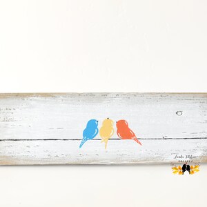 May include: A rectangular piece of weathered white wood featuring a horizontal black line with three painted birds perched on it. The birds are colored blue, yellow, and orange from left to right. The text Linda Fehlor Gallery is visible.