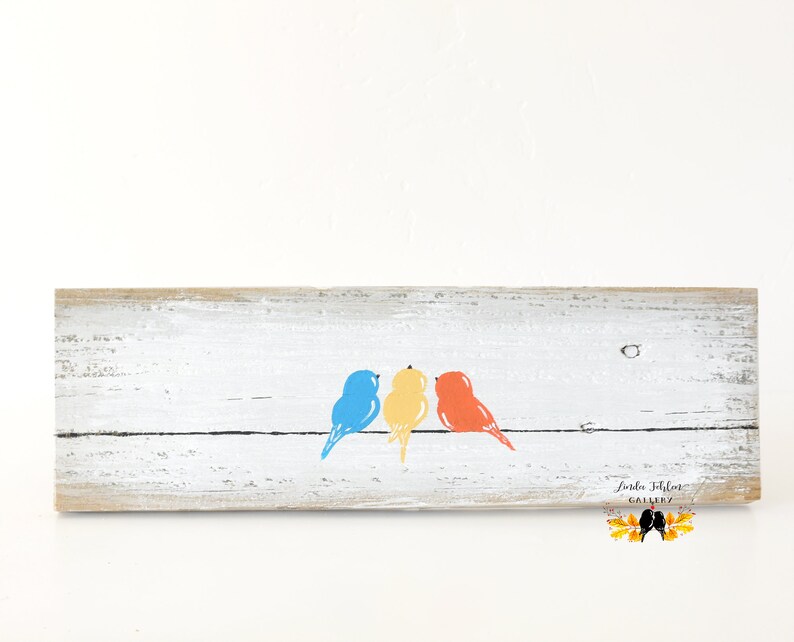 May include: A rectangular piece of weathered white wood featuring a horizontal black line with three painted birds perched on it. The birds are colored blue, yellow, and orange from left to right. The text Linda Fehlor Gallery is visible.