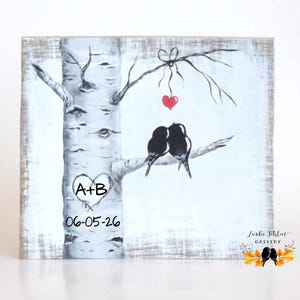 May include: A square wooden sign featuring a painted birch tree with two black birds perched on a branch. A red heart hangs from a twig above the birds. The tree trunk displays a carved heart with the text A+B and the date 06-05-26 below it.