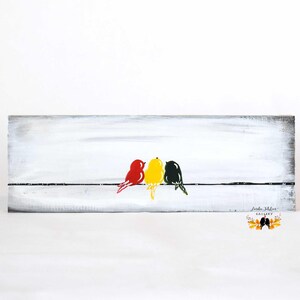 rustic wood board painted with white background with three birds on a wire. bird on left is red, middle bird is yellow, and right bird is dark green.