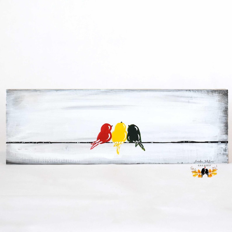 rustic wood board painted with white background with three birds on a wire. bird on left is red, middle bird is yellow, and right bird is dark green.