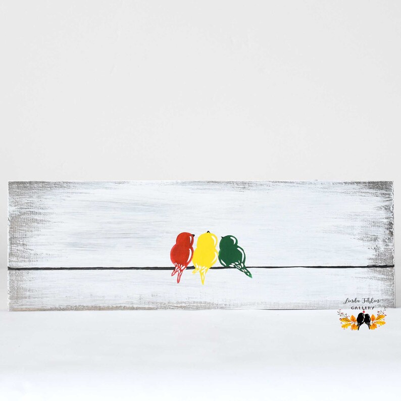 May include: A rectangular wooden sign with a distressed white finish featuring a thin black horizontal line. Three stylized birds in red, yellow, and green are perched on the line. The text Linda Fehlner Gallery is printed in the bottom right corner.