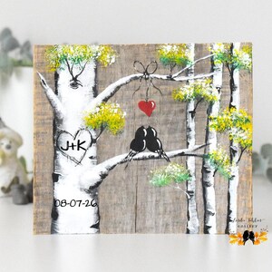 May include: A rectangular wooden plaque featuring a hand-painted scene of white birch trees with green foliage. Two black birds sit on a branch below a red heart hanging from a bow. A heart carved with J+K and the date 08-07-26 appear on the trunk.