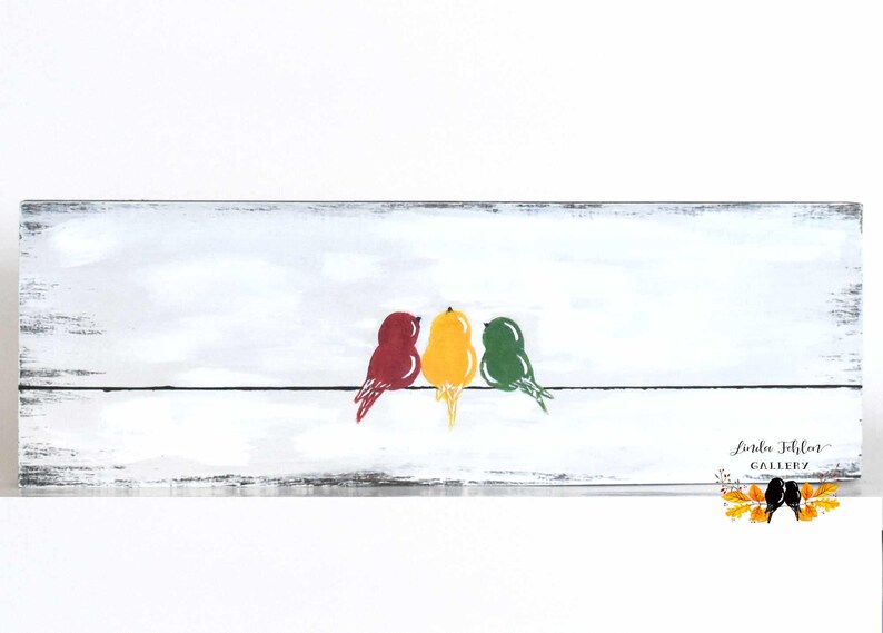 May include: A rectangular wooden sign with a distressed white finish featuring a painted black horizontal line with three birds perched on it. The birds are colored red, yellow, and green. The text Linda Fehlor GALLERY is printed in the bottom right corner.
