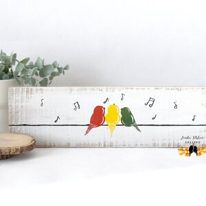 May include: A rectangular white wooden sign with a distressed finish featuring a horizontal black line with three birds in red, yellow, and green perched on it. Black musical notes are scattered above the birds, and the text Linda Folsom GALLERY is printed in the bottom right corner.