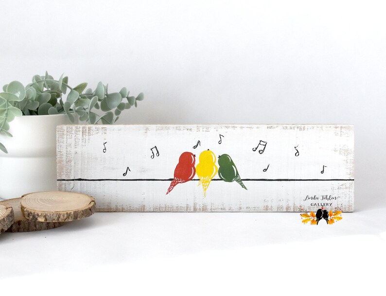 May include: A rectangular white wooden sign with a distressed finish featuring a horizontal black line with three birds in red, yellow, and green perched on it. Black musical notes are scattered above the birds, and the text Linda Folsom GALLERY is printed in the bottom right corner.