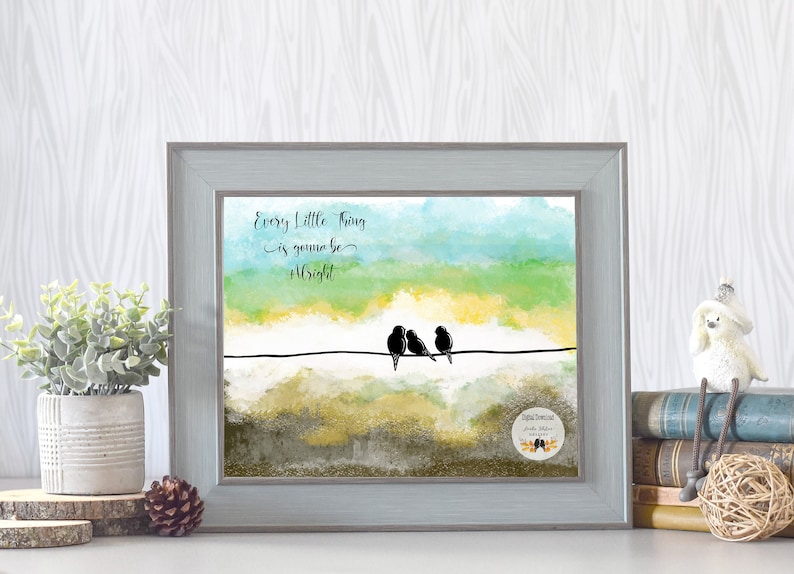 May include: A watercolor painting of a sunset with three birds perched on a wire. The text "Every little thing is gonna be alright" is written in black cursive above the birds. The painting is framed in a light gray frame.