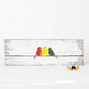 May include: A rectangular white distressed wooden sign featuring a horizontal black line with three stylized birds perched on it. The birds are colored red, yellow, and green from left to right. The text Linda Tehlon Gallery is printed in the corner.