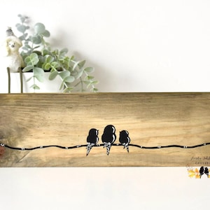 May include: A rectangular piece of light brown wood featuring a painted black line across the center with three black bird silhouettes perched on it. The bottom right corner displays the text "Lierdee Schlon GALLERY" in a cursive font.