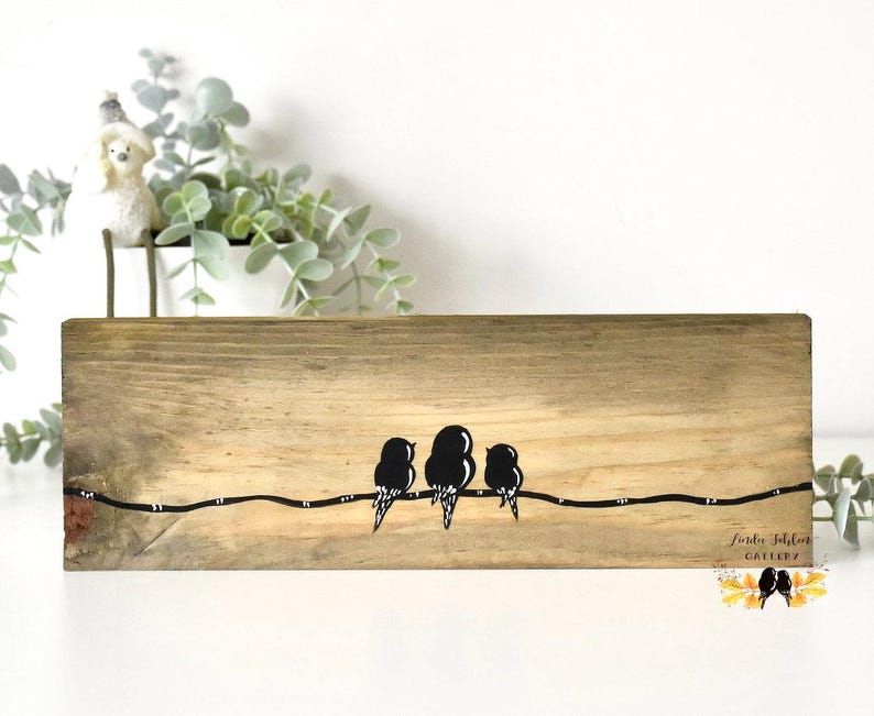 May include: A rectangular piece of light brown wood featuring a painted black line across the center with three black bird silhouettes perched on it. The bottom right corner displays the text "Lierdee Schlon GALLERY" in a cursive font.