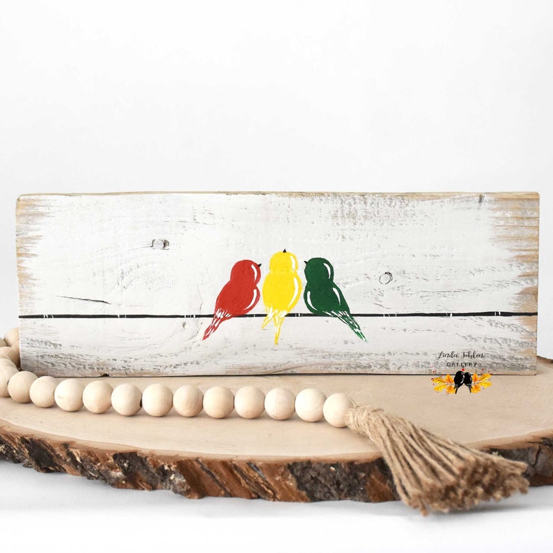May include: A rectangular piece of distressed white wood featuring a painted black horizontal line with three birds perched on it. The birds are colored red, yellow, and green from left to right. The text "Linda Johler Gallery" is printed in black.