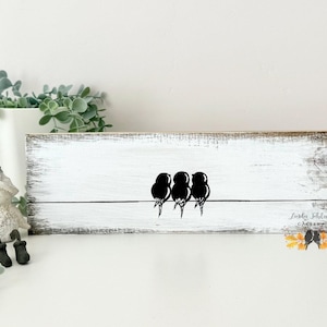 May include: A rectangular wooden sign with a distressed white finish featuring a black graphic of three birds perched on a single horizontal line. The bottom right corner displays the text Linda Schlez Gallery with a small colorful leaf graphic.