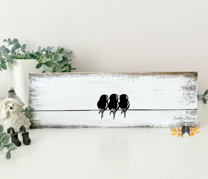 May include: A rectangular wooden sign with a distressed white finish featuring a black graphic of three birds perched on a single horizontal line. The bottom right corner displays the text Linda Schlez Gallery with a small colorful leaf graphic.