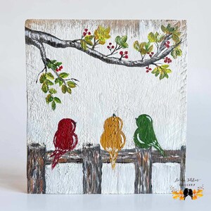 May include: A rectangular wooden plaque featuring a hand-painted scene of three birds in red, yellow, and green perched on a brown fence. Above the birds, a tree branch with green leaves and red berries extends across the white-washed wood surface.
