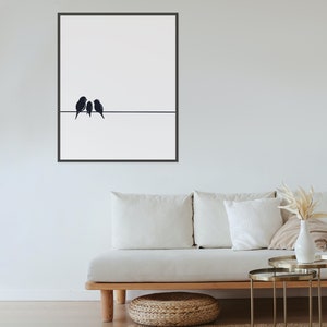 May include: A black and white minimalist art print featuring three birds perched on a wire. The print is framed in a black frame and hangs on a white wall above a white couch.