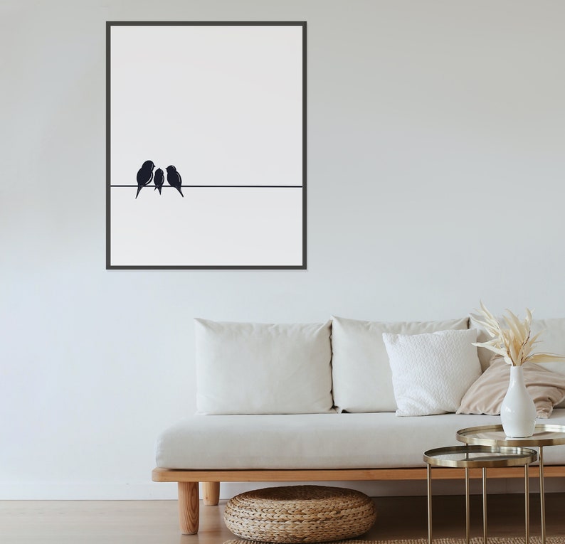 May include: A black and white minimalist art print featuring three birds perched on a wire. The print is framed in a black frame and hangs on a white wall above a white couch.