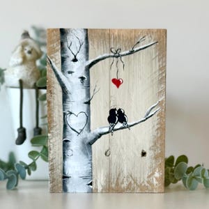 May include: A rectangular wooden plaque featuring a hand-painted white birch tree on a light brown wood grain background. The tree trunk displays a carved heart outline, while two black birds sit on a branch below a single red heart hanging by a bow.