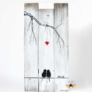 May include: A white wooden sign with a black and white drawing of a tree branch with a red heart hanging from it. Two black birds are perched on a wire below the branch. The sign also includes the text "M+K" and "08-19-24".