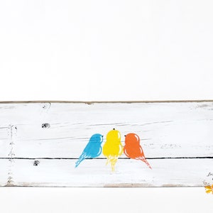 May include: A rectangular white-painted wooden board featuring a horizontal black line with three stylized birds perched on it. The birds are colored blue, yellow, and orange from left to right. The bottom right corner displays the text Linda Fohler Gallery.