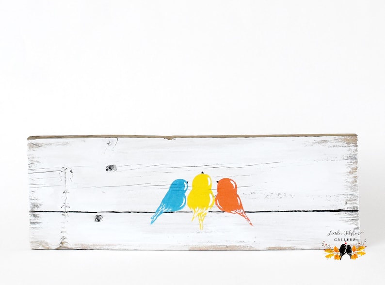 May include: A rectangular white-painted wooden board featuring a horizontal black line with three stylized birds perched on it. The birds are colored blue, yellow, and orange from left to right. The bottom right corner displays the text Linda Fohler Gallery.