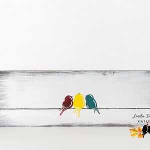 May include: A rectangular wooden sign with a distressed white finish featuring a black horizontal line with three stylized birds in maroon, yellow, and teal. The bottom right corner displays the text Linda Fehlen GALLERY with a leaf graphic.
